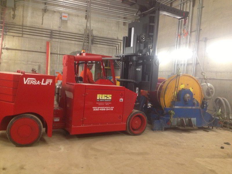 Red Versa-Lift forklift moving heavy industrial equipment in a concrete-walled facility with visible pipes and wires.