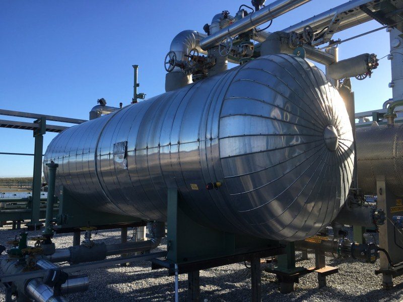 Large industrial metal tank with pipes and valves, set in an outdoor facility under a clear blue sky.