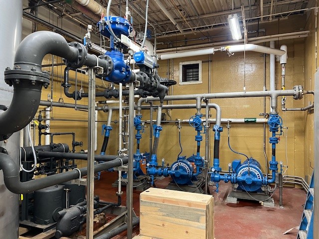 Industrial mechanical room with blue pumps, pipes, and valves. Yellow walls and overhead lighting visible.