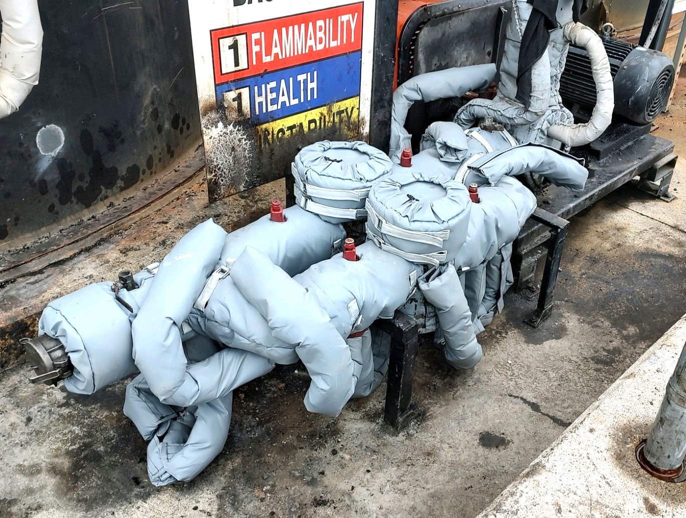 Insulated industrial equipment with multiple pipes is placed on a concrete floor, next to a flammability and health sign.